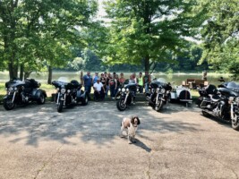A group of people stands behind five parked motorcycles near a lake, with a dog in the foreground.